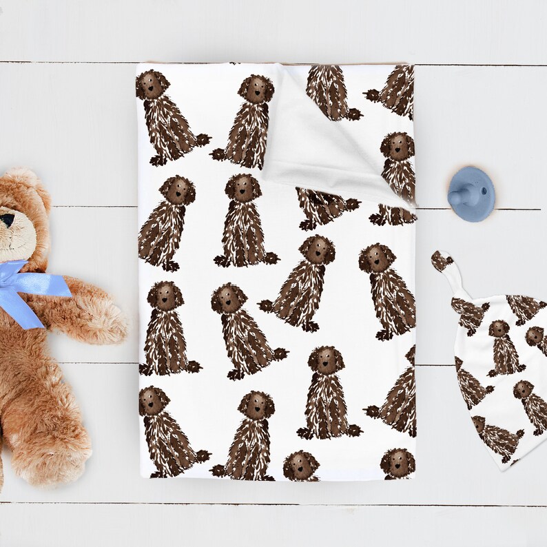 May include: A white baby blanket featuring a repeating pattern of brown, shaggy-haired dogs. The blanket is folded at the top corner, revealing a plain white underside. The fabric has a soft texture and a consistent print of the canine illustrations.