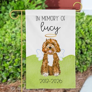 Pode incluir: Uma bandeira de jardim branca com uma ilustração em aguarela de um cão castanho e branco com asas de anjo e um halo dourado. O texto diz IN MEMORY OF lucy e 2013-2026. A bandeira é exibida num suporte de metal dourado.