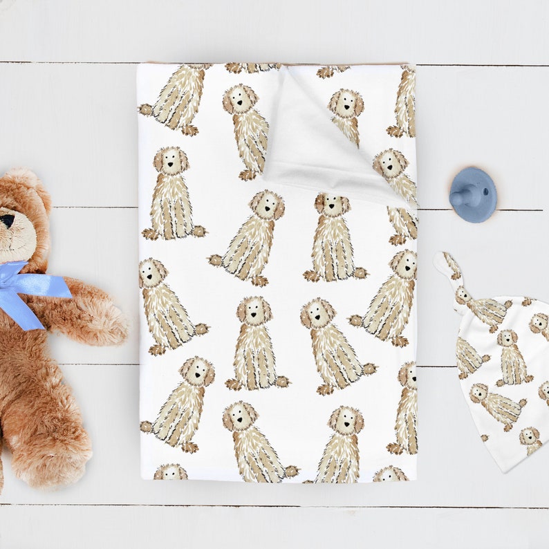 May include: A white baby blanket and matching hat featuring a repeating pattern of illustrated tan, shaggy-haired dogs. The blanket is folded at the top corner, revealing a plain white underside. A brown teddy bear with a blue ribbon is nearby.