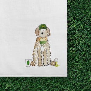 May include: A white tea towel featuring a watercolor illustration of a fluffy tan dog wearing a green cap and a green lanyard with a white tag. The dog is positioned between a white cup with a green oval and a white golf ball on a yellow tee.