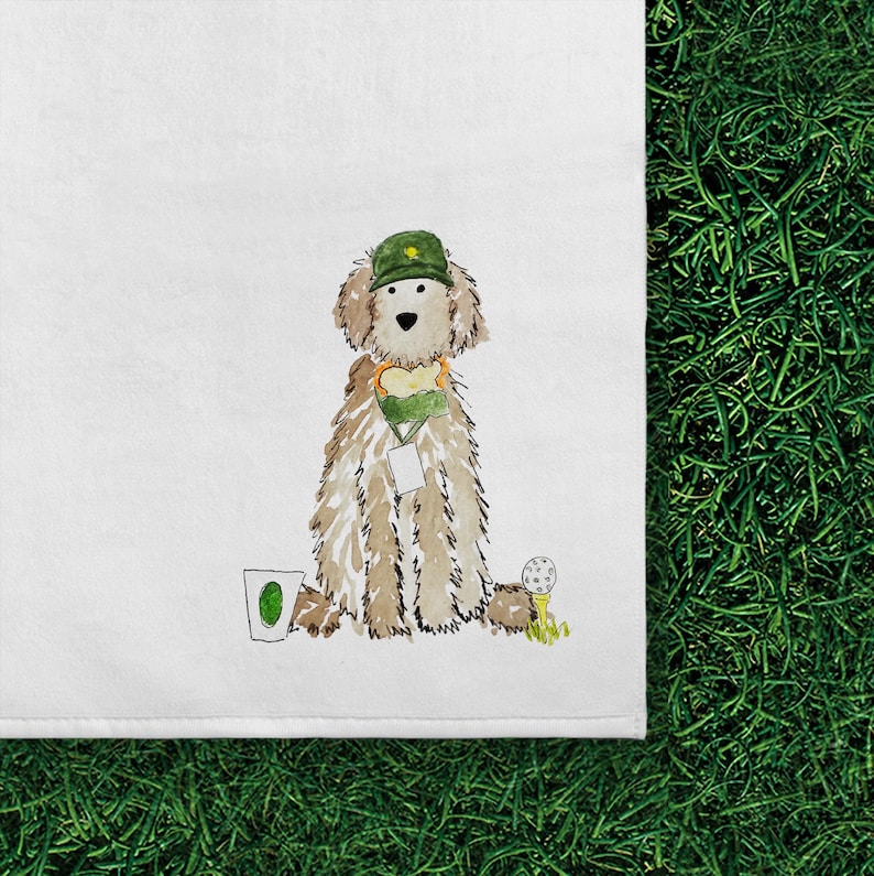 May include: A white tea towel featuring a watercolor illustration of a fluffy tan dog wearing a green cap and a green lanyard with a white tag. The dog is positioned between a white cup with a green oval and a white golf ball on a yellow tee.