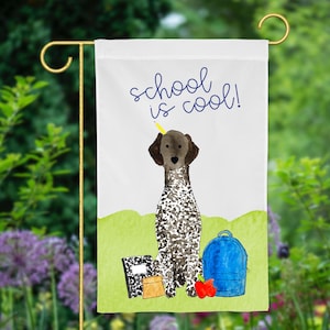 May include: A white garden flag with a brown and white dog sitting in front of a blue backpack, a red apple, and a notebook. The text "school is cool!" is written in blue on the flag.