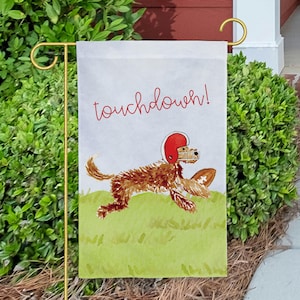 May include: A rectangular garden flag featuring a light gray background with the red cursive text touchdown! above a brown doodle dog wearing a red football helmet and running across a light green grassy field while holding a brown football.
