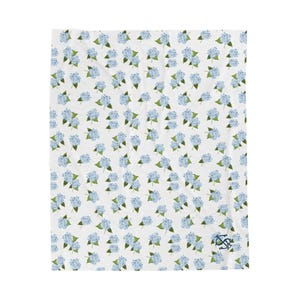 Può includere: Coperta bianca con un motivo ripetuto di fiori di ortensia blu e foglie verdi. Un piccolo logo ricamato si trova nell'angolo in basso a destra. Il design è floreale e la coperta è probabilmente per l'arredamento della casa.