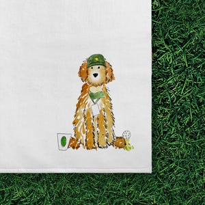 May include: A white tea towel featuring a watercolor illustration of a golden-colored dog wearing a green golf cap and a green scarf with a white tag. To the left is a white cup with a green oval, and to the right is a white golf ball on a tee.