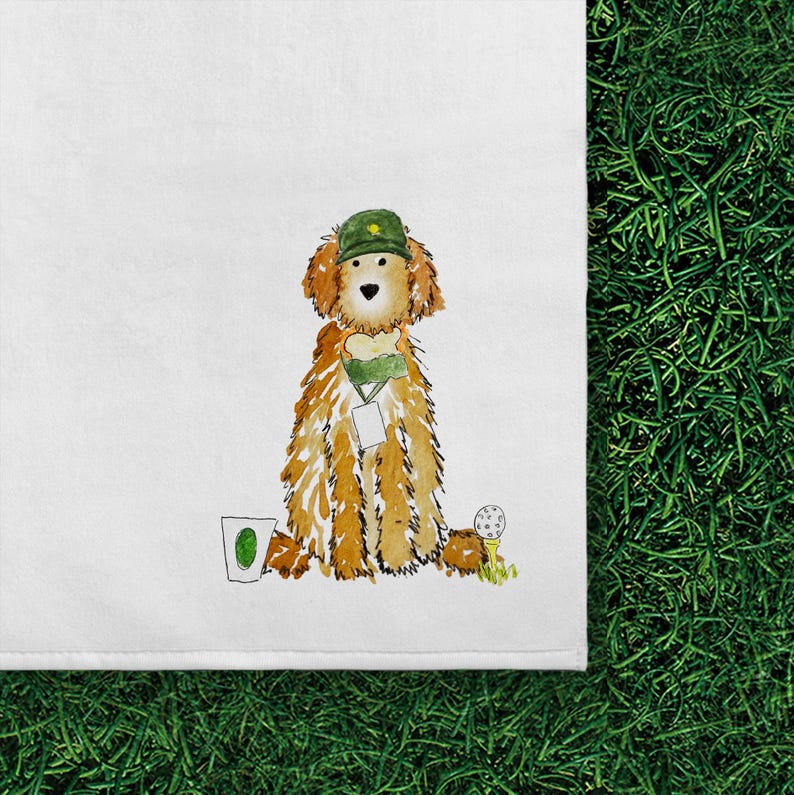 May include: A white tea towel featuring a watercolor illustration of a golden-colored dog wearing a green golf cap and a green scarf with a white tag. To the left is a white cup with a green oval, and to the right is a white golf ball on a tee.