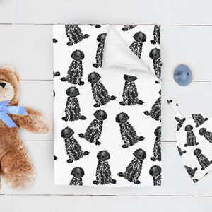 May include: A white rectangular blanket featuring a repeating black and white illustration of a fluffy dog, a matching white baby hat with the same dog pattern, a brown plush teddy bear with a blue ribbon, and a light blue pacifier.