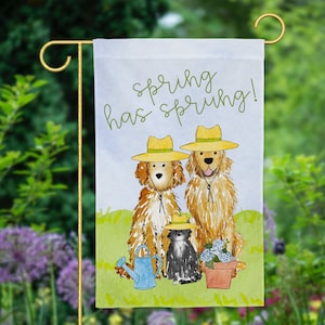May include: A white garden flag with the text "spring has sprung!" in black cursive writing. Two golden retrievers wearing straw hats are sitting in front of a black cat wearing a straw hat. A blue watering can and a pot of purple flowers are in the foreground.