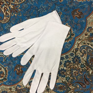 Puede incluir: Un par de guantes de algodón blanco. Los guantes están sobre una tela con estampado azul y dorado.
