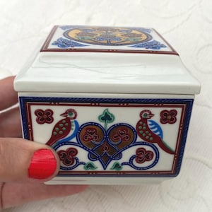 May include: A rectangular white ceramic box with a lid, featuring a colourful decorative pattern of two birds facing a central heart-shaped motif in shades of red, blue, green, and gold, framed by thin dark blue and red border lines.