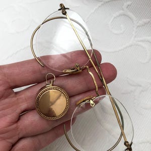 May include: Antique gold-toned eyeglasses with round lenses and a matching round pendant. The eyeglasses feature delicate wire frames and a bridge. The pendant has a beaded border and a loop for hanging. The items are displayed against a white background.