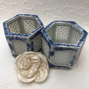 Può includere: Due vasi esagonali in porcellana bianca con bordatura floreale blu e lati traforati. Un fiore di stoffa color crema con accenti di perle è posto davanti ai vasi. I vasi sono probabilmente per scopi decorativi.