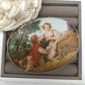 May include: Oval-shaped antique trinket box with a painted scene of a couple in a pastoral setting. The box has a gold-colored rim and is set in a gray jewelry box. A white satin flower with pearl accents is in the background.