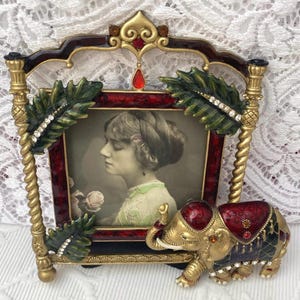 May include: A rectangular picture frame featuring a deep red enamel border, gold-tone metal accents, and green leaf motifs with clear crystals. A gold-tone elephant figurine with red and black enamel and crystal accents is attached to the base.
