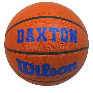 May include: A textured orange basketball with black rubber seams. The ball features the blue text DAXTON printed in a bold serif font above the blue Wilson logo. Below the logo, the text INDOOR GAME BALL is printed in small black letters.