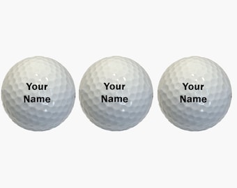 May include: Three white golf balls arranged in a horizontal row. Each ball features a dimpled surface and the black text "Your Name" printed clearly in the center. The design is consistent across all three items, showcasing a standard spherical shape.