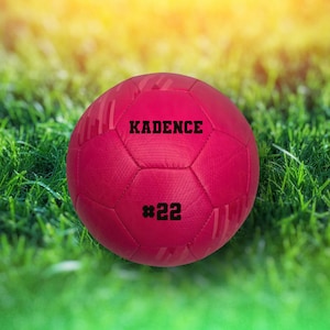 May include: A pink soccer ball with black stitching and the name "KADENCE #22" printed on it.