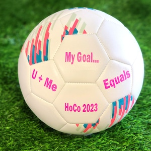 May include: White soccer ball with pink and teal stripes. The ball has the text "My Goal... U + Me Equals HoCo 2023" printed on it.