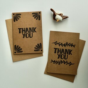 May include: Two brown thank you cards with black ink. One card has a simple "Thank You" message with a decorative border. The other card has a "Thank You" message with a floral border.