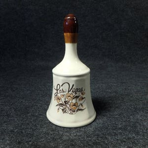 May include: White ceramic bell with a brown top and a floral design. The bell says "Las Vegas" on the front.