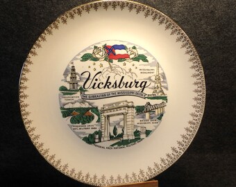 May include: A white ceramic plate with a gold rim and a colorful illustration of Vicksburg, Mississippi. The illustration includes the text "Vicksburg, The Gibraltar of the Mississippi Delta".
