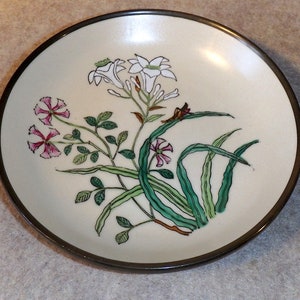 May include: A round, off-white ceramic dish with a dark brown rim. The dish features a floral design with white and pink flowers, green leaves, and stems. The design is centered on the dish.