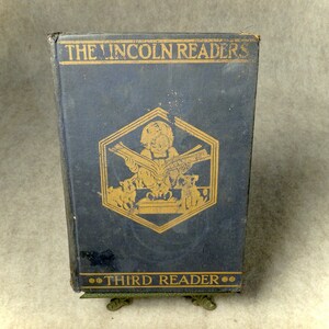May include: A vintage, dark blue hardcover book titled "The Lincoln Readers: Third Reader". The cover features a gold illustration of a child reading a book with two dogs at their feet.