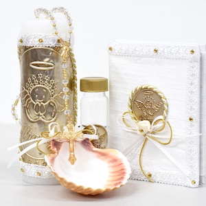 May include: A white baptism set with a white candle, a small glass bottle, a seashell, and a white booklet with gold accents. The candle has a gold angel design and a white lace trim. The booklet has a gold angel seal and a white ribbon.