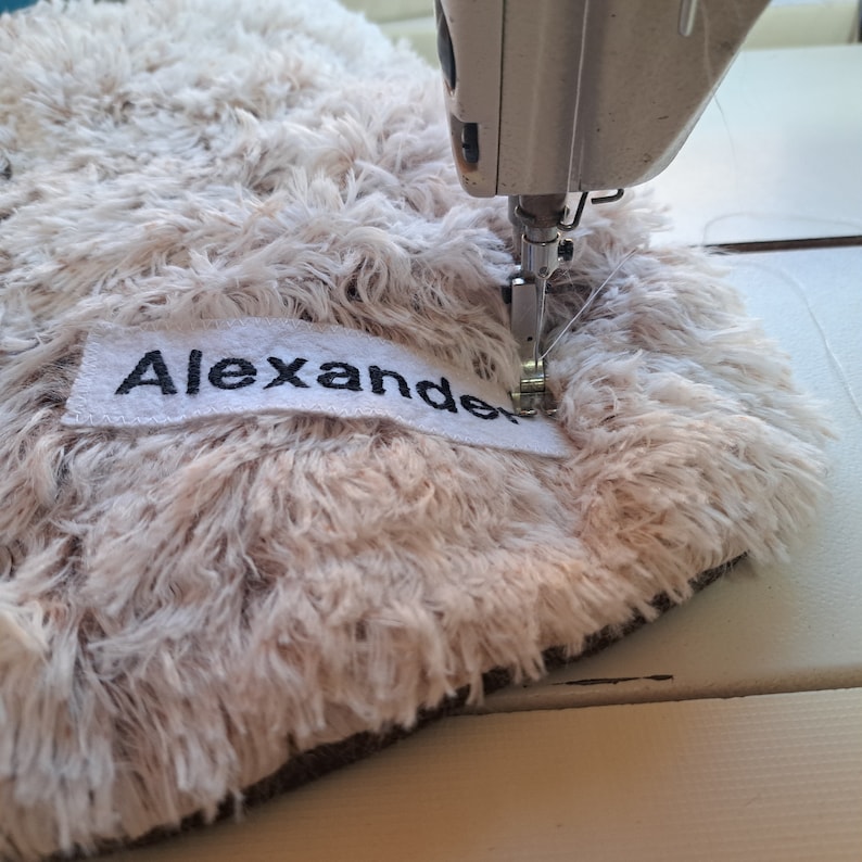 Close-up of a sewing machine embroidering the name 'Alexander' on a plush fabric, highlighting our personalized customization service for unique, bespoke nursery decor.