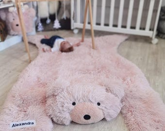 May include: A plush, pink bear-shaped rug with a soft, furry texture. The rug features a bear face with black eyes and a black nose. The name "Alexandra" is printed on a white label on the rug. A wooden baby gym is set up above the rug.