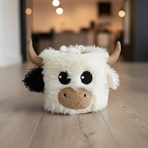 A white stuffed cow toy with brown spots and horns sits on a wooden floor.