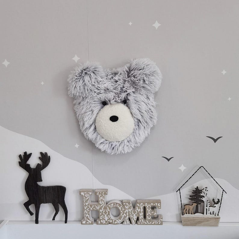 a wall decorated with a large stuffed bear head, a wooden deer, and a wooden house with a birdhouse. The bear head is mounted on the wall, while the wooden deer and house are placed below it. The background of the image is a light gray color with white stars scattered across it.