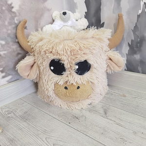 A plush toy cow with large horns and a teddy bear sitting on top of it, placed on a wooden floor against a patterned wall.