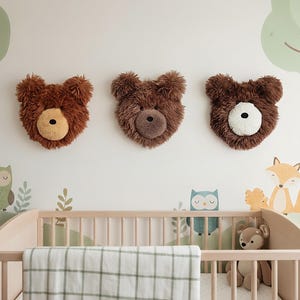 Könnte beinhalten: Drei Wanddekorationen in Form von Teddybärköpfen mit braunem Fell und beigen oder weißen Akzenten sind an einer weißen Wand angebracht. Unter den Dekorationen befindet sich ein Kinderbett mit einer karierten Decke und Stofftieren.