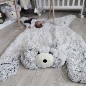 Könnte beinhalten: Ein flauschiger, grau-weißer Bärenteppich mit weißer Schnauze und schwarzen Augen. Der Teppich hat ausgestreckte Arme und liegt auf einem hellen Holzboden. Der Name "Alexander" ist auf einer der Bärenpfoten aufgedruckt.