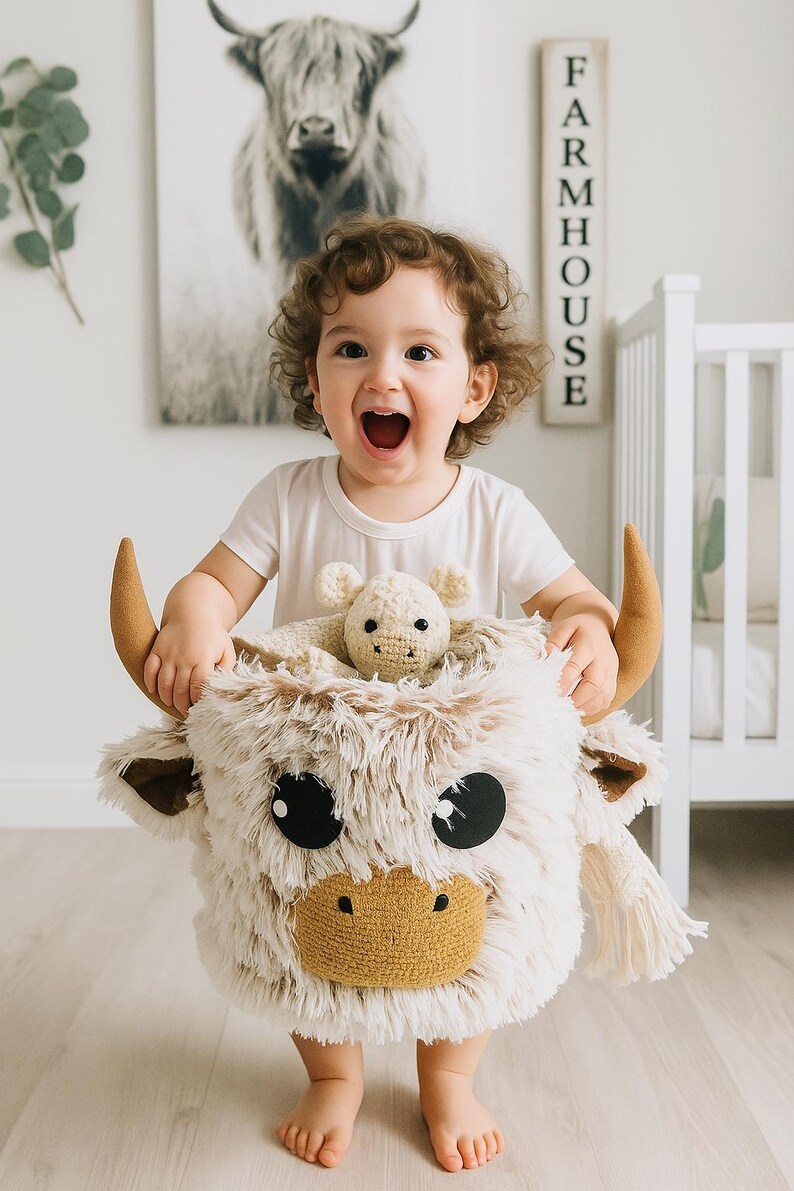 Handmade Highland Cow storage basket crafted from soft Shaggy Minky fleece in creamy beige and tan, featuring 3D plush horns, floppy ears, a textured brown snout, and oversized black felt eyes. Sized approx. 10 x 10 x 9 inches, this lightweight and flexible basket is perfect for holding toys, books, or baby essentials. A playful and functional nursery accessory that adds warmth and personality to farm animal or Scandi-boho themed spaces.