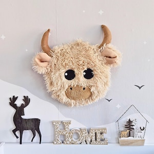 May include: A fluffy, light brown Highland cow head wall decoration with large black eyes, tan horns, and ears. The cow is mounted on a white wall with decorative elements, including a wooden deer silhouette and the word "HOME".
