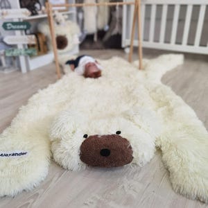 A large white plush polar bear toy is lying on a wooden floor, with a baby sleeping on top of it.