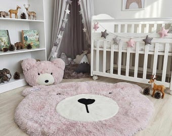 May include: A plush, pink bear-shaped rug with a white face and black nose. A matching bear head pillow is next to the rug. The nursery setting includes a white crib, star garland, and a framed bear illustration.