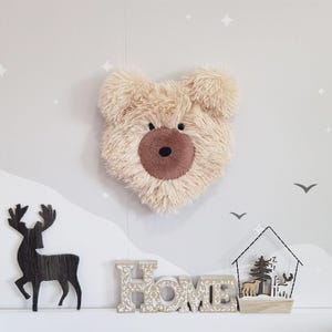 May include: A fluffy, beige bear head wall decoration with a brown nose and inner ears. The bear is mounted on a light grey wall with a decorative deer silhouette, the word "HOME" in wooden letters, and a small house ornament.