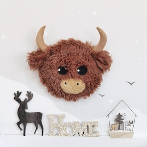 May include: A fluffy, brown Highland cow head with big black eyes and tan horns. The cow is mounted on a white wall with decorative elements, including a wooden deer silhouette and the word "HOME".