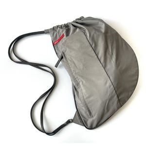 May include: Gray drawstring backpack with a front zippered pocket and black drawstrings. A small red rectangular tag is visible on the upper right side. The bag is made of a lightweight, possibly nylon, material.