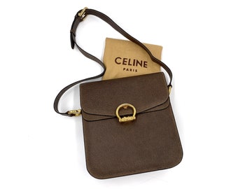 Op de afbeelding: Bruine leren crossbodytas met een goudkleurige ring sluiting. De tas heeft een klepsluiting en een lange, verstelbare riem. Een beige stoffen tas met de tekst "CELINE PARIS" staat achter de tas.