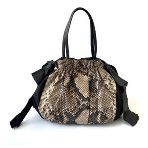 Peut inclure: Un sac à main noir et à motif peau de serpent avec des poignées et des accents noirs. Le sac est doté d'une fermeture à cordon et de la marque "PRADA" en lettres dorées. Le motif peau de serpent est dans les tons marron, beige et noir.