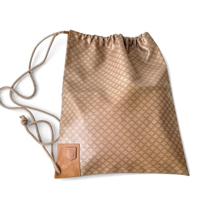Puede incluir: Mochila con cordón beige con un patrón repetitivo. La bolsa presenta un parche de cuero marrón con un logotipo. El cordón es una cuerda marrón claro. La bolsa está hecha de un material brillante.