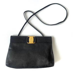 May include: A black shoulder bag with a textured surface, featuring a horizontal fabric band across the top. A gold-toned metal hardware piece with the embossed text Salvatore Ferragamo is centered on the band. It has a thin black shoulder strap.