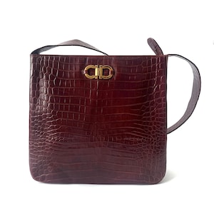 May include: A deep burgundy shoulder bag with a textured crocodile-embossed pattern. The bag features a square silhouette and a gold-toned metal Gancini clasp centered on the front. It includes a matching leather shoulder strap attached to the sides.