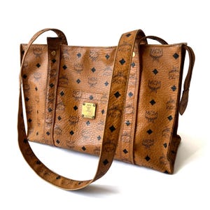 Peut inclure: Sac fourre-tout en cuir marron avec un motif répété de losanges noirs et le logo MCM. Le sac est doté de deux bandoulières et d'une petite plaque rectangulaire dorée. Le sac est de taille moyenne.