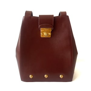 May include: A structured dark burgundy leather backpack featuring a gold-tone metal clasp closure with the Salvatore Ferragamo logo. The bag includes two shoulder straps and three decorative gold-tone metal studs along the bottom edge.
