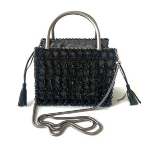 May include: A black handbag featuring a structured square silhouette with a woven grid overlay. It includes a silver-toned metal top handle, a matching silver snake chain shoulder strap, and two side drawstrings finished with black tassels.